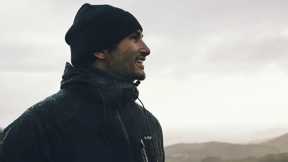 lächelnder Wanderer mit Mütze und schwarzer Jacke vor Bergen unter einem diesigen Himmel